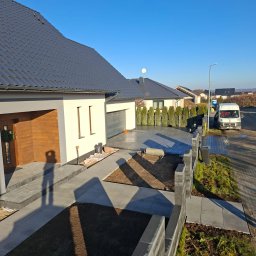 DaMaR Usługi koparką - Nowoczesny dom z szarej kostki brukowej i częściowo ukończonym ogrodzeniem z betonowych bloczków. Widoczny cień osoby robiącej zdjęcie. Słoneczny dzień.