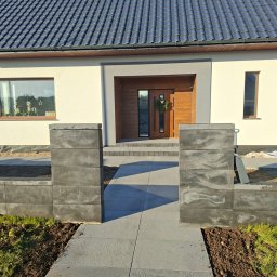 DaMaR Usługi koparką - Nowoczesny dom jednorodzinny z szarą kostką brukową prowadzącą do wejścia. Widoczne elementy ogrodzenia z betonowych bloczków i świeżo obsadzona ziemia.