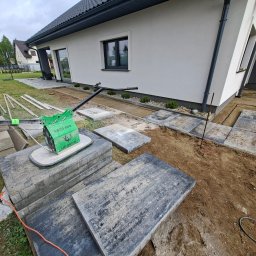 Koparka i Kostka -prace ziemne Koszalin - Układanie płyt chodnikowych w ogrodzie przy domu. Widoczny ubijak wibracyjny, płyty z szarego betonu i wytyczone ścieżki. Prace w toku, częściowo ułożona nawierzchnia.