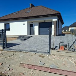 Koparka i Kostka -prace ziemne Koszalin - Nowoczesny dom jednorodzinny z podjazdem wyłożonym szarą kostką brukową. Widoczny fragment ogrodzenia i zagęszczarka. Słoneczny dzień, błękitne niebo.