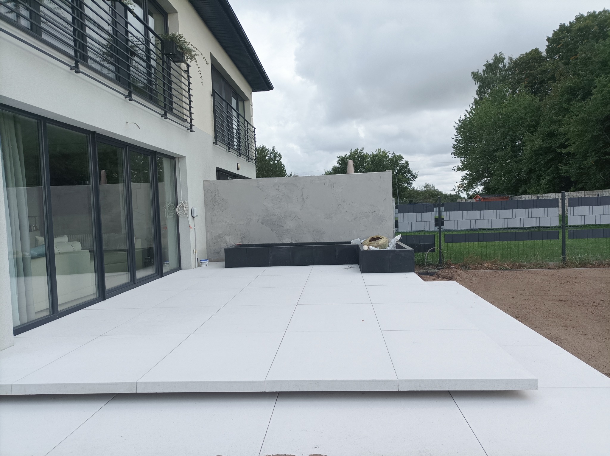 Nowoczesny taras z jasnych płyt, z czarną donicą i widokiem na ogród. Minimalistyczny design, duże przeszklenia domu i stalowe balustrady.