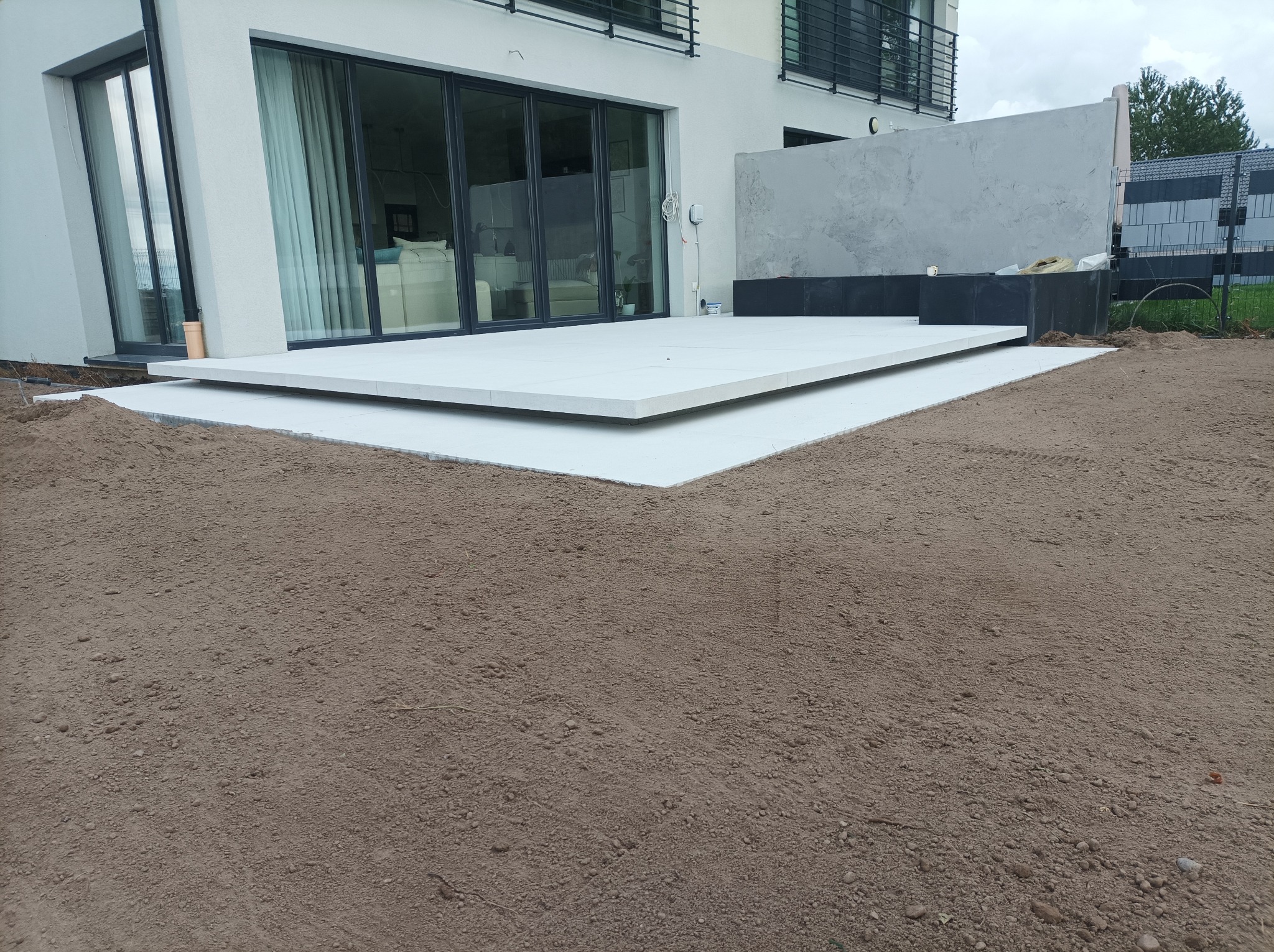 Nowoczesny taras z jasnych płyt na przygotowanym podłożu z ziemi ogrodowej, przylegający do budynku z dużymi oknami przesuwnymi i minimalistycznym ogrodzeniem.