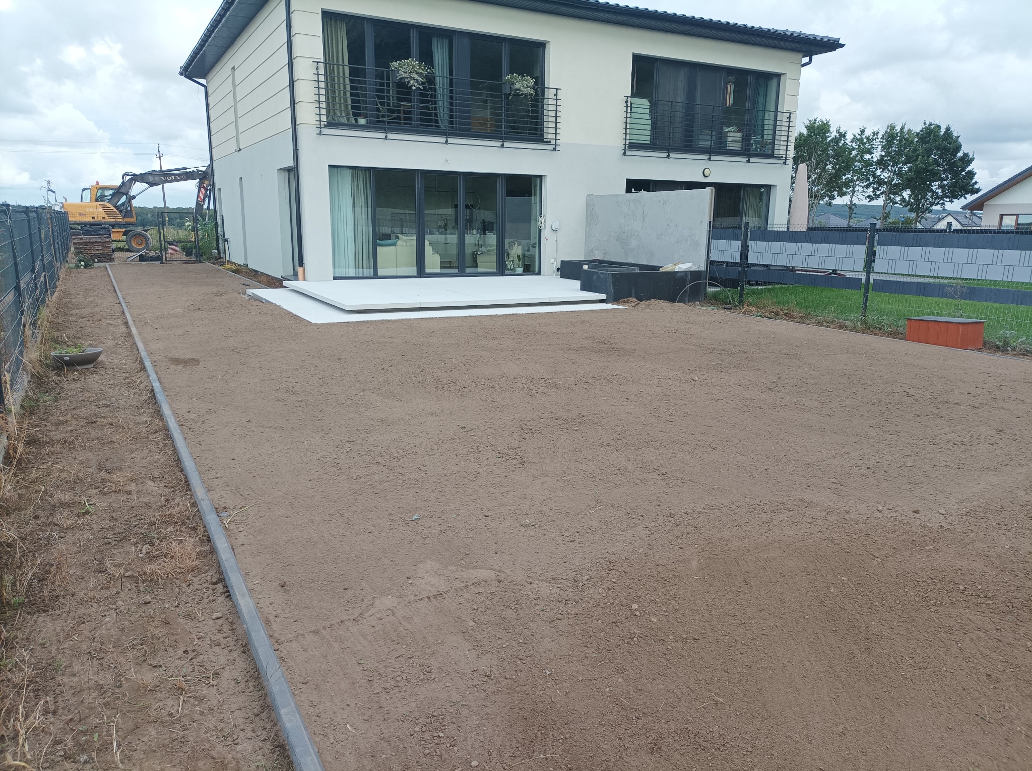 Przygotowany teren pod trawnik z widocznym domem, obrzeżami i koparką w tle. Wyrównana ziemia, gotowa pod zasiew lub rozłożenie trawy z rolki.