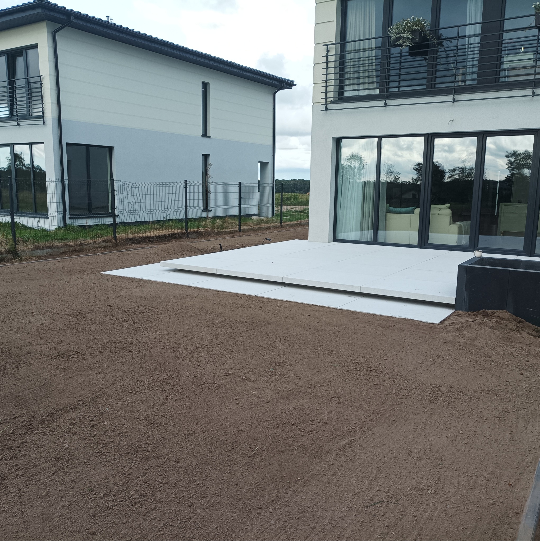 Nowoczesny taras z jasnych płyt, ziemia przygotowana pod trawnik, widok na dwa budynki mieszkalne i ogrodzenie z siatki.