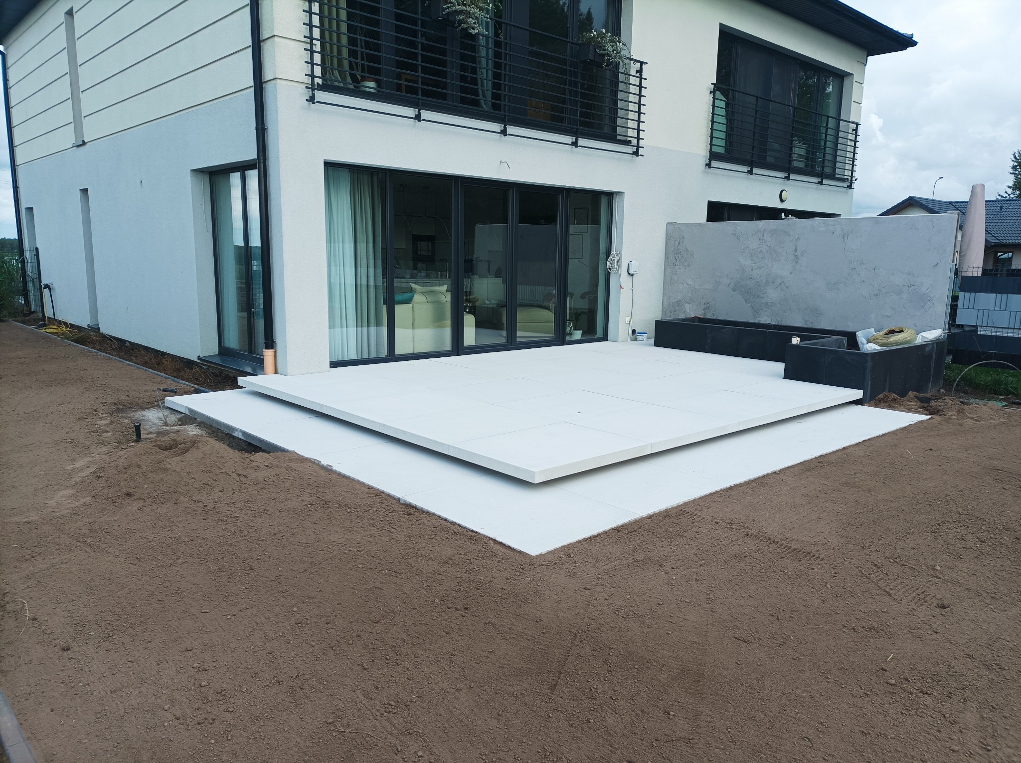 Nowoczesny taras z jasnych płyt na przygotowanym podłożu, widok na budynek z dużymi oknami i balkonem z czarną balustradą, ziemia ogrodowa w tle.