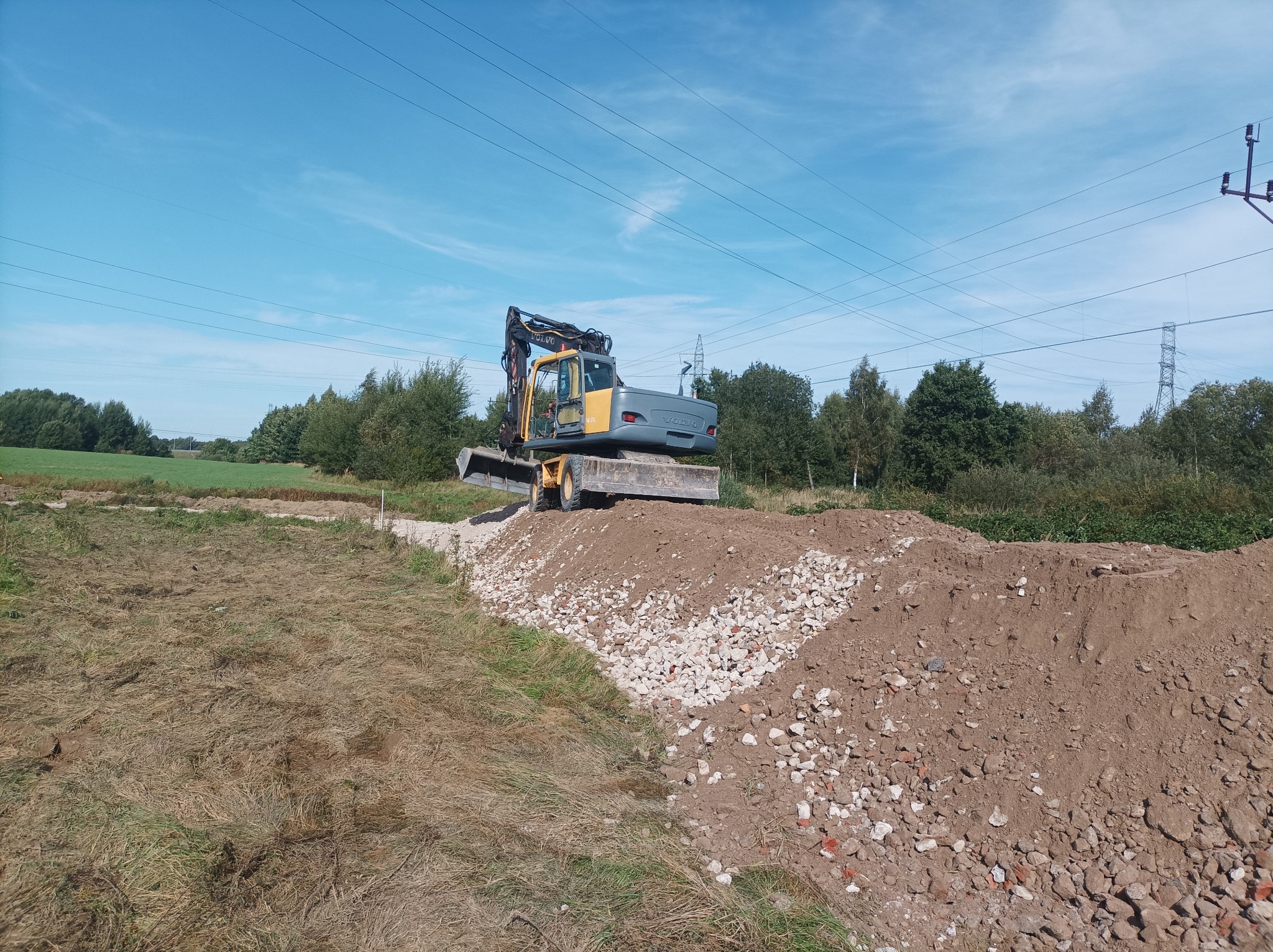 Żółto-szara koparka Volvo na wzgórzu z gruzem i ziemią. W tle błękitne niebo, drzewa i słupy wysokiego napięcia. Prace ziemne w trakcie realizacji.