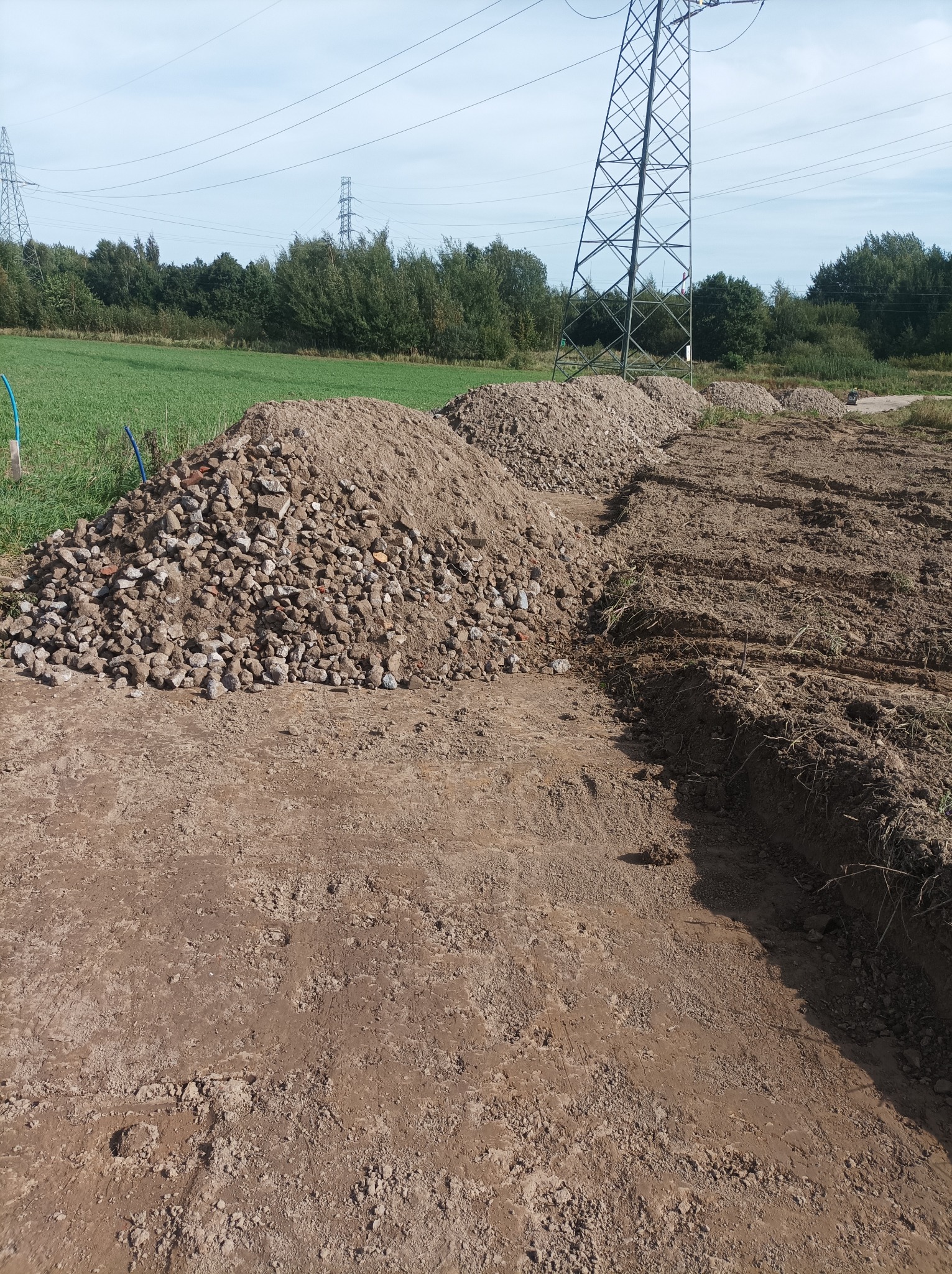 Przygotowanie terenu pod budowę: wyrównany grunt, pryzmy gruzu i ziemi, słupy wysokiego napięcia w tle. Widok z poziomu gruntu.
