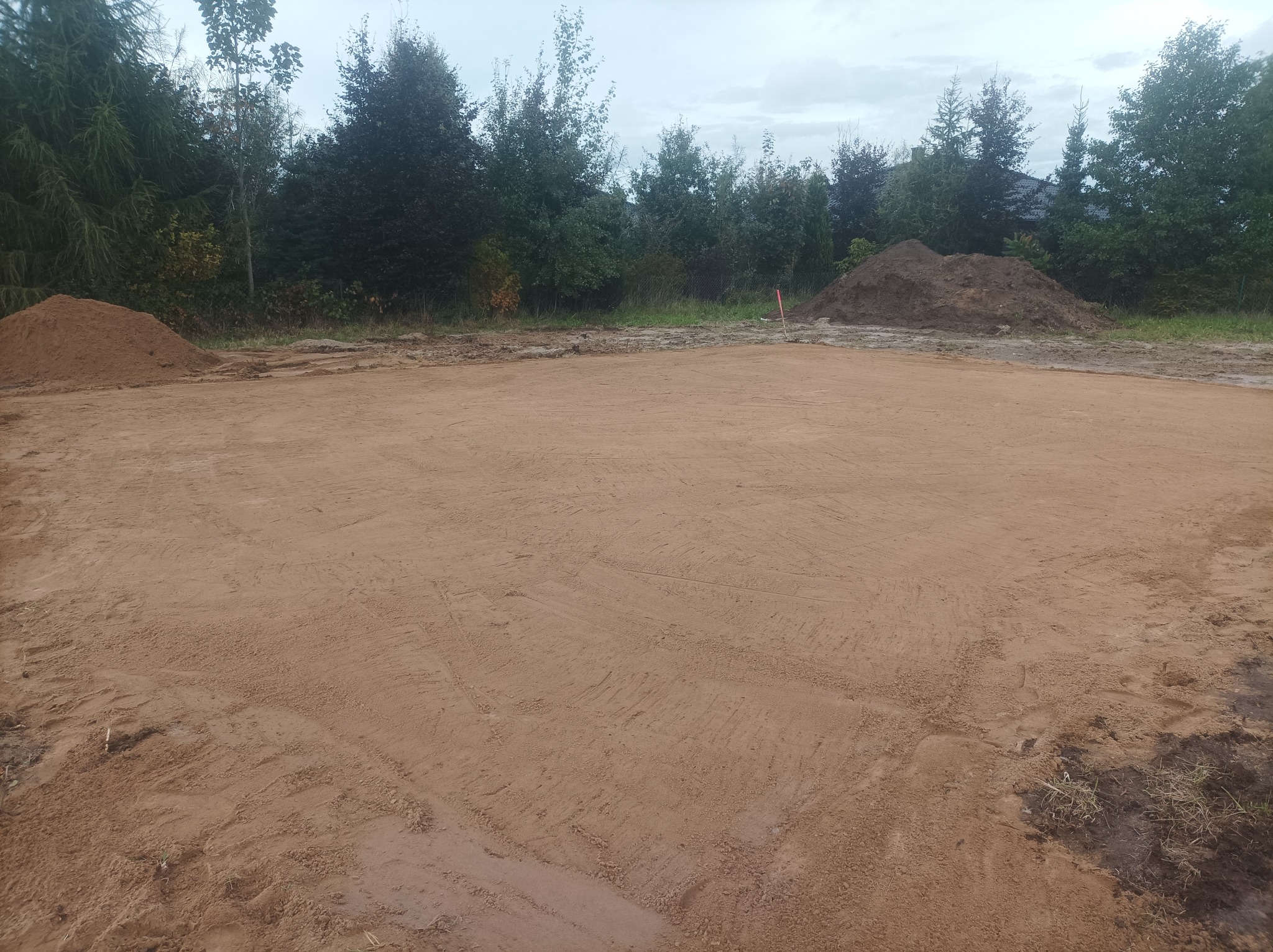 Wyrównany, piaszczysty teren pod budowę, z hałdami ziemi w tle i zielenią drzew. Widoczne ślady maszyn niwelujących grunt. Pochmurne niebo.