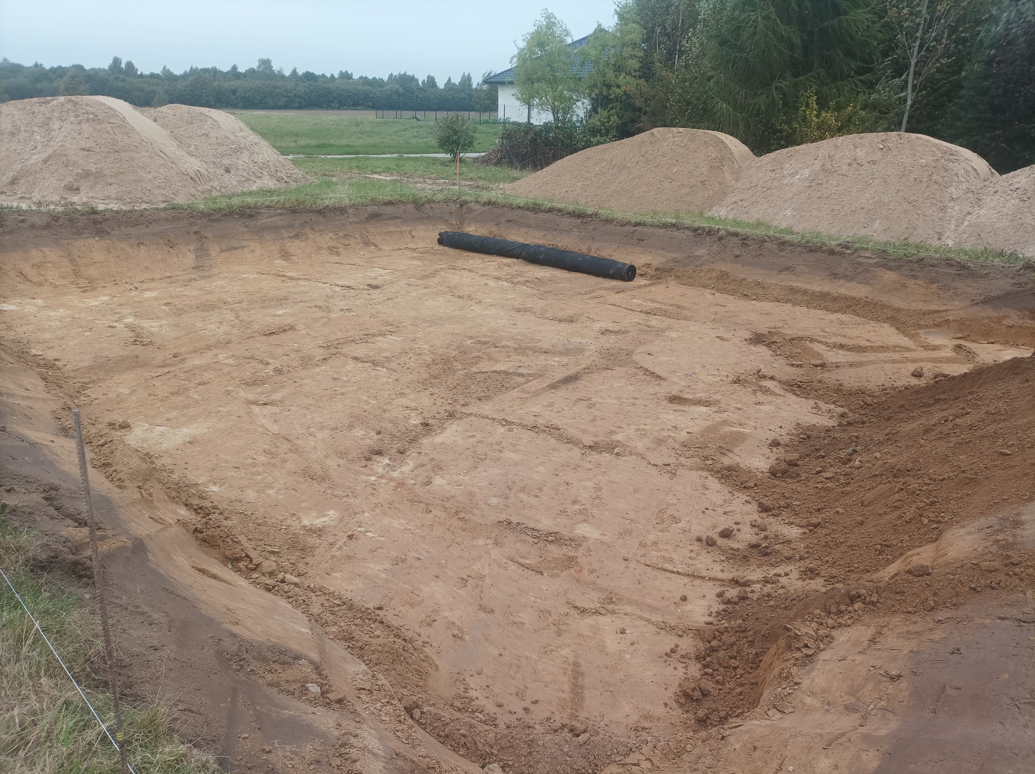 Wyrobisko ziemne przygotowane pod budowę stawu, z wałami ziemi wokół i czarną geowłókniną. W tle widoczny dom i drzewa.