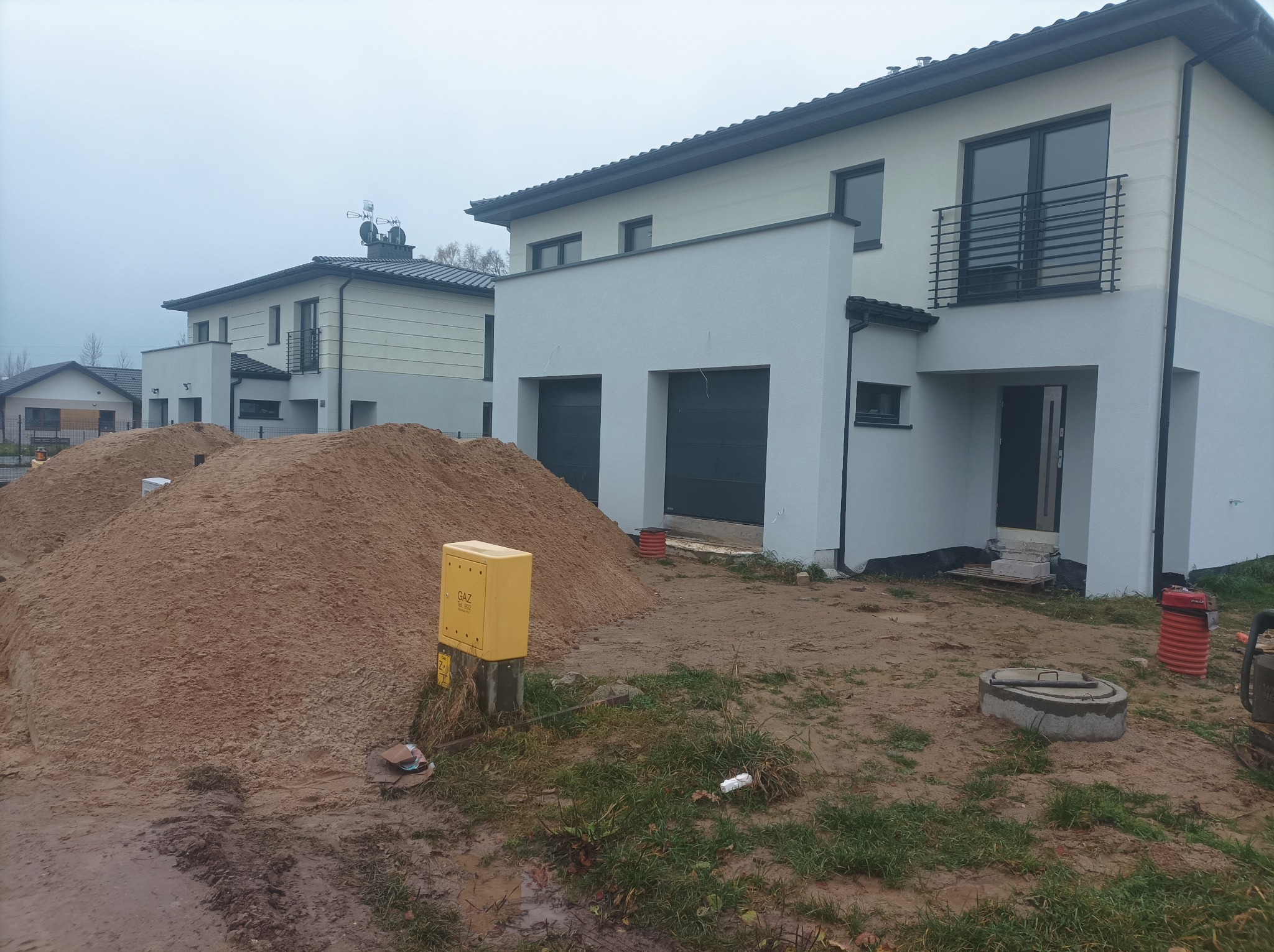 Nowoczesny dom jednorodzinny w trakcie budowy, z widoczną stertą piasku i studzienką kanalizacyjną na działce. Szara, pochmurna pogoda. Żółta skrzynka gazowa.