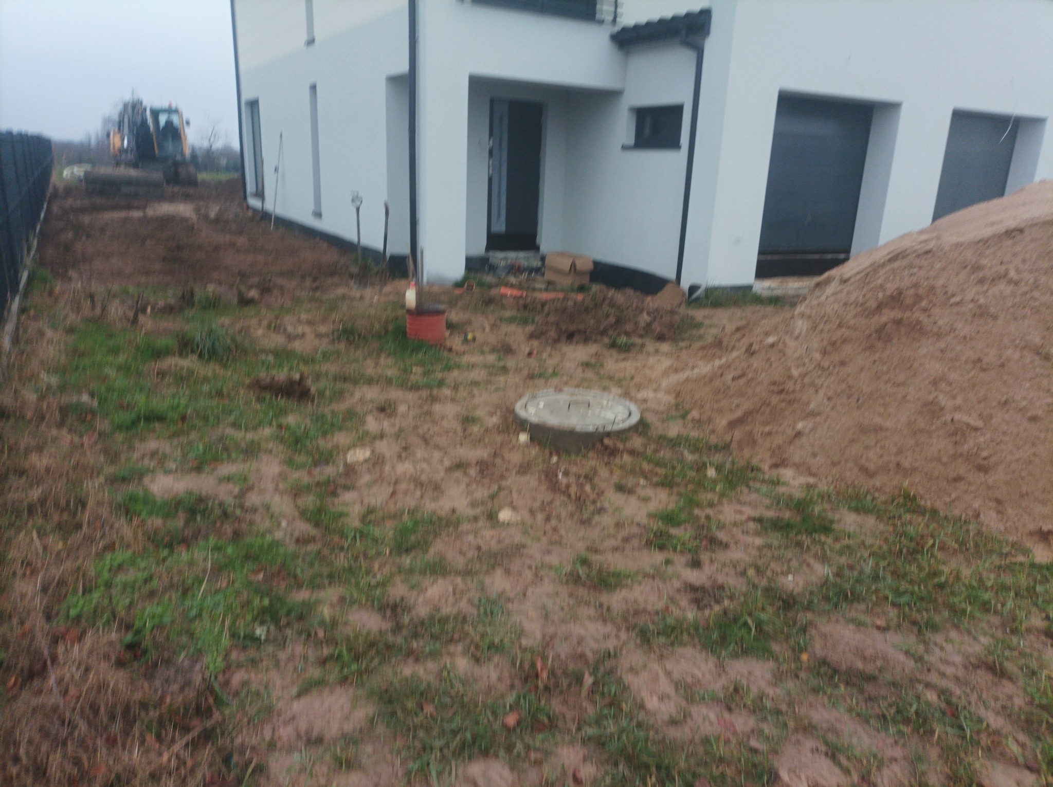 Nowy dom w trakcie budowy: wykopane fundamenty, hałda piasku, widoczny ciężki sprzęt. Szara, pochmurna pogoda. W tle ogrodzenie.