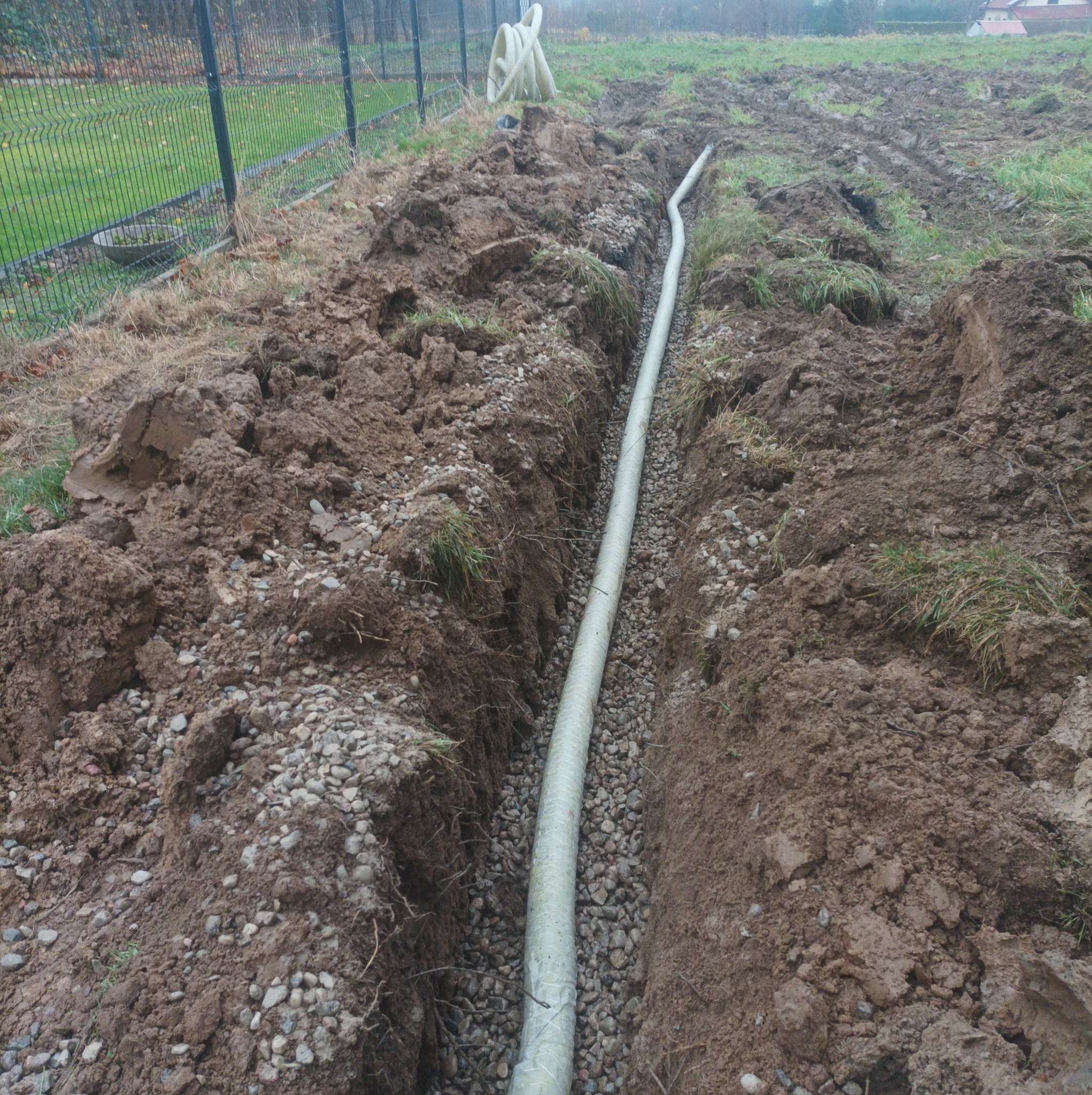 Biała rura w wykopie wypełnionym żwirem, widok z góry. W tle ziemia, trawa i ogrodzenie z siatki. Prace ziemne, instalacja podziemna.