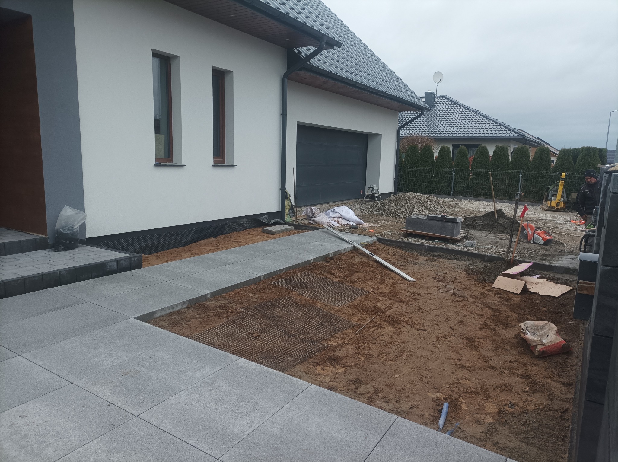 Układanie szarej kostki brukowej na ścieżce przed nowoczesnym domem z szarą dachówką. Widoczne narzędzia i materiały budowlane. Prace w toku, częściowo wykończona nawierzchnia.