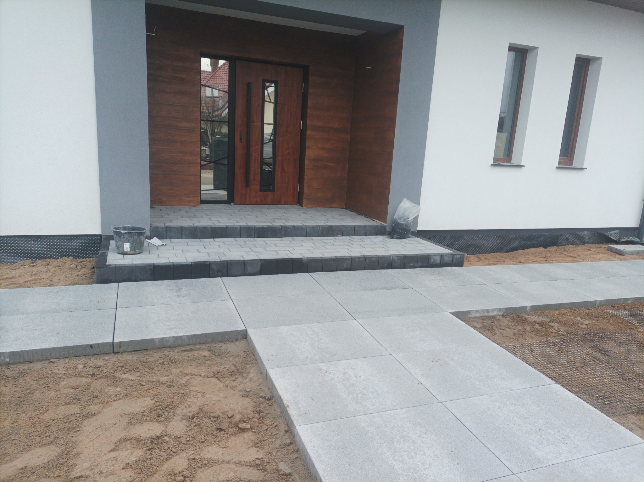 Nowoczesne wejście do domu z szarej kostki brukowej. Ułożony chodnik prowadzi do drewnianych drzwi wejściowych. Widoczne elementy elewacji i przygotowany teren.