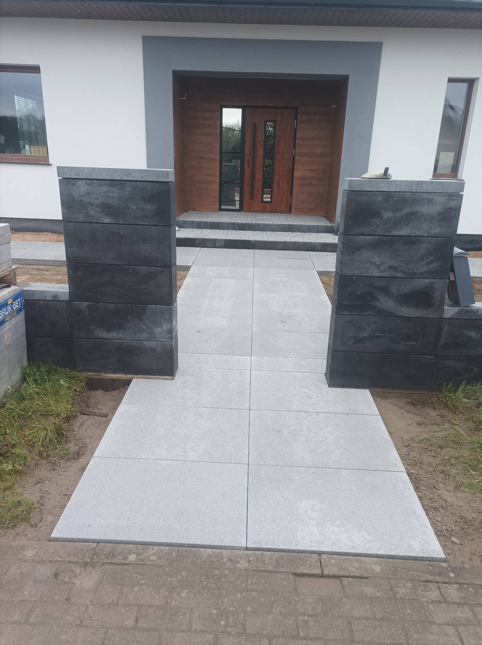 Nowoczesna ścieżka z szarych płyt prowadząca do domu, flankowana przez ciemne, modułowe słupki. Widoczne detale wykończenia i paleta z kostką brukową.