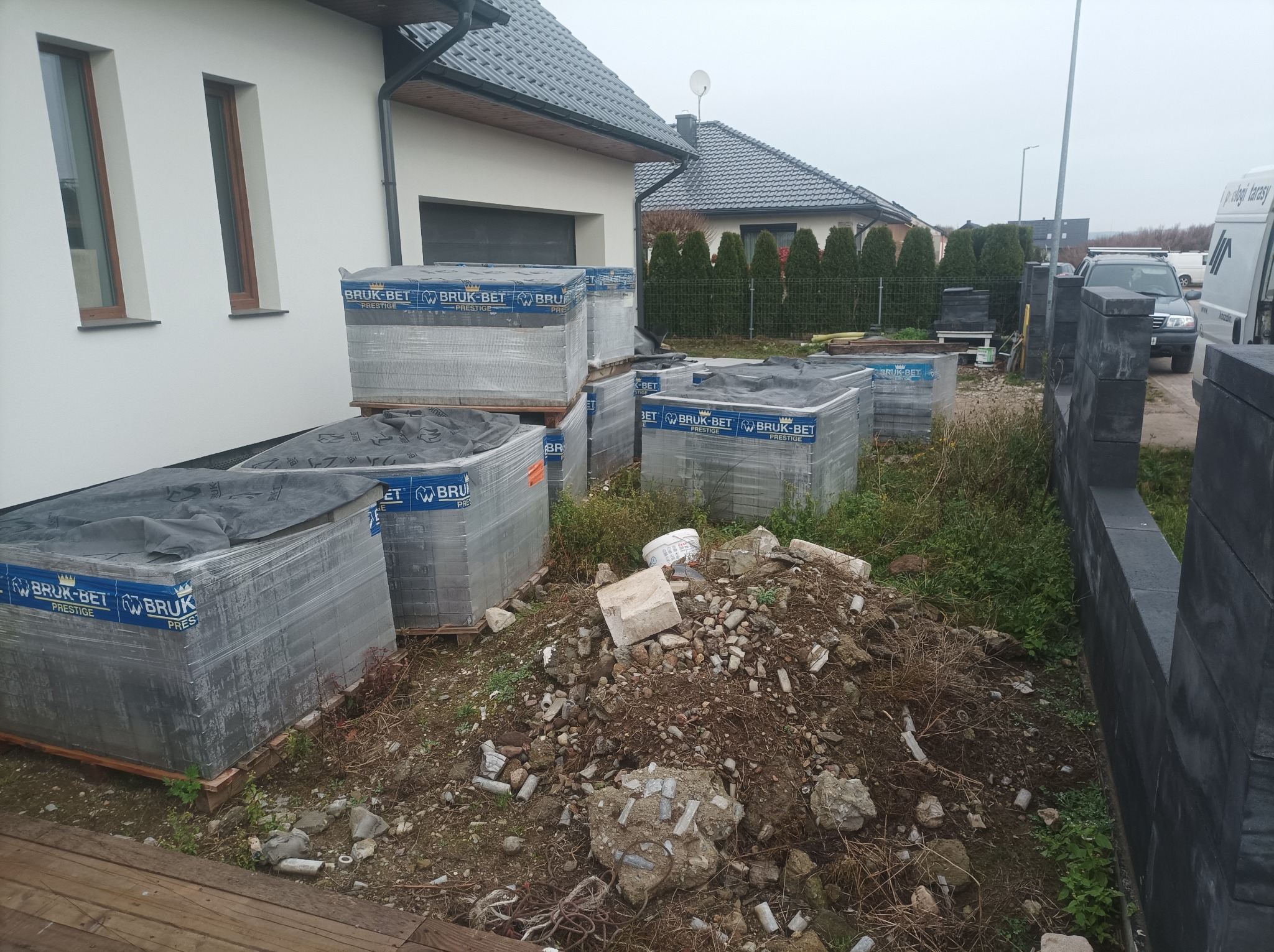 Palety kostki brukowej Bruk-Bet Prestige obok domu z szarą dachówką i fragmentem czarnego ogrodzenia. Na pierwszym planie gruz i ziemia.