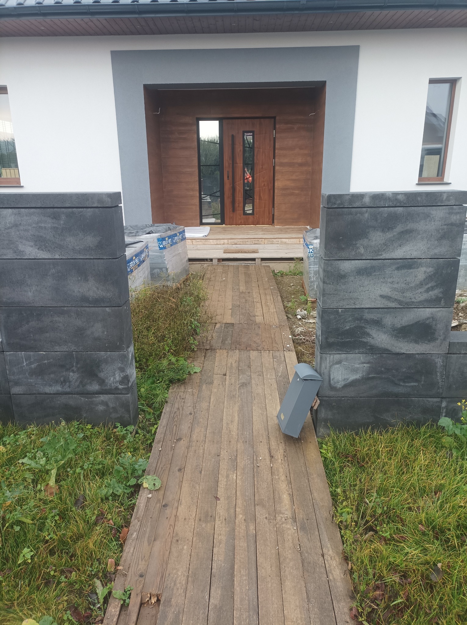 Wejście do domu z drewnianą ścieżką i elementami ogrodzenia z szarej kostki. Widoczne materiały budowlane i skrzynka na listy. Ujęcie z perspektywy.