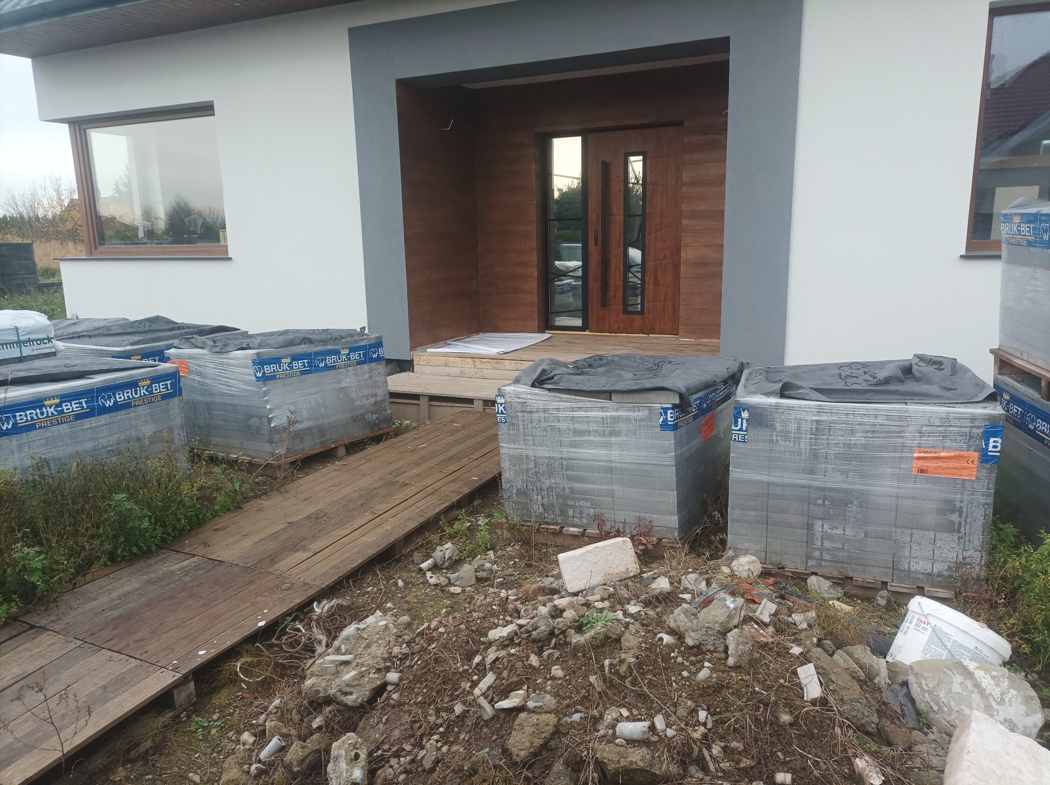 Nowoczesny dom z paletami kostki brukowej Bruk-Bet Prestige przed wejściem. Przygotowanie do układania nawierzchni, widoczny drewniany chodnik i gruz.