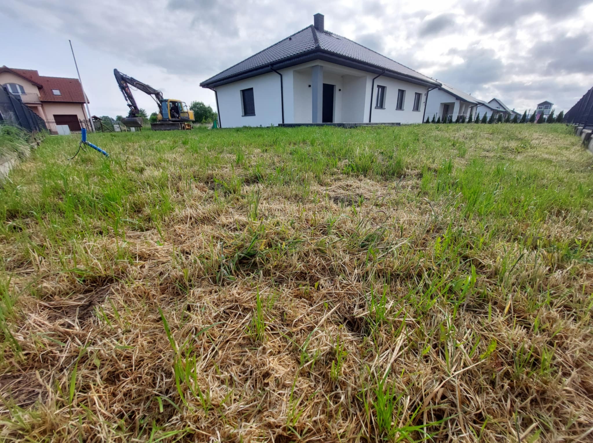 Nowoczesny dom jednorodzinny z ciemnym dachem i jasną elewacją, z widocznym na trawniku sprzętem budowlanym. W tle sąsiednie domy i pochmurne niebo.