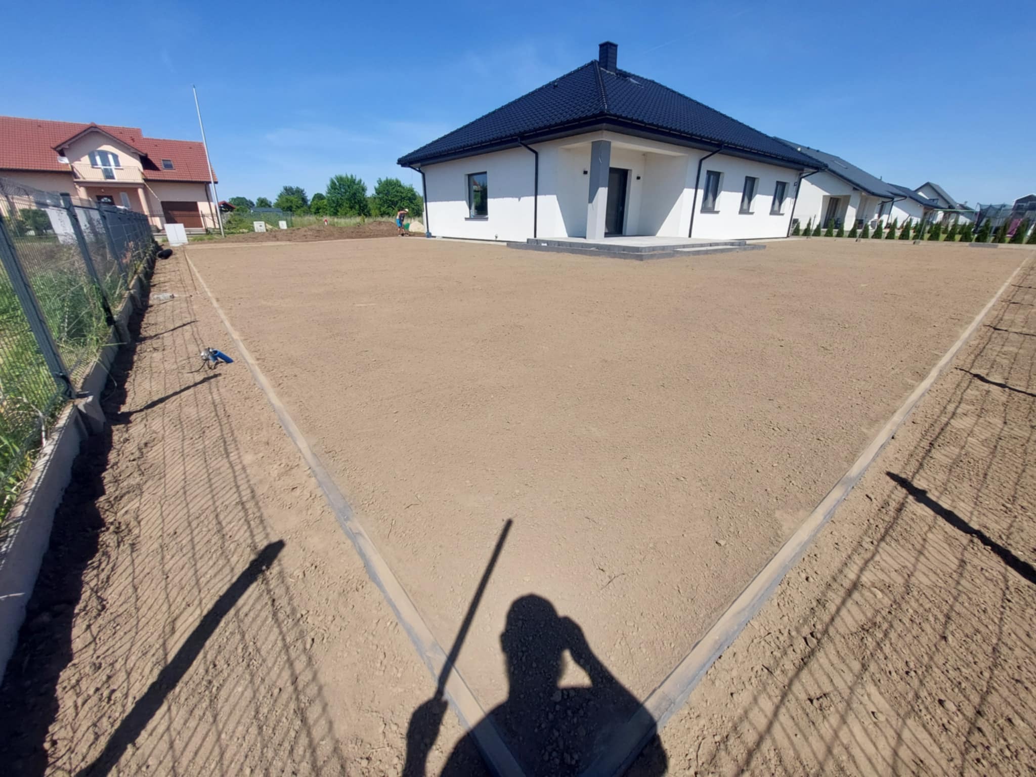 Przygotowany teren pod trawnik z obrzeżami, widok na nowoczesny dom z ciemnym dachem i ogrodzenie siatkowe. Czyste niebo, cień osoby w rogu kadru.