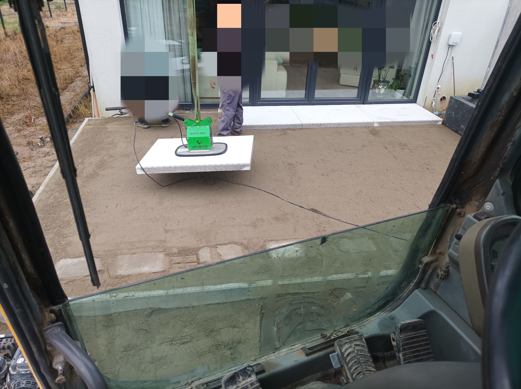 Układanie płyt chodnikowych na piasku przy użyciu specjalistycznego urządzenia w pobliżu domu z dużymi oknami, widok z kabiny maszyny budowlanej.