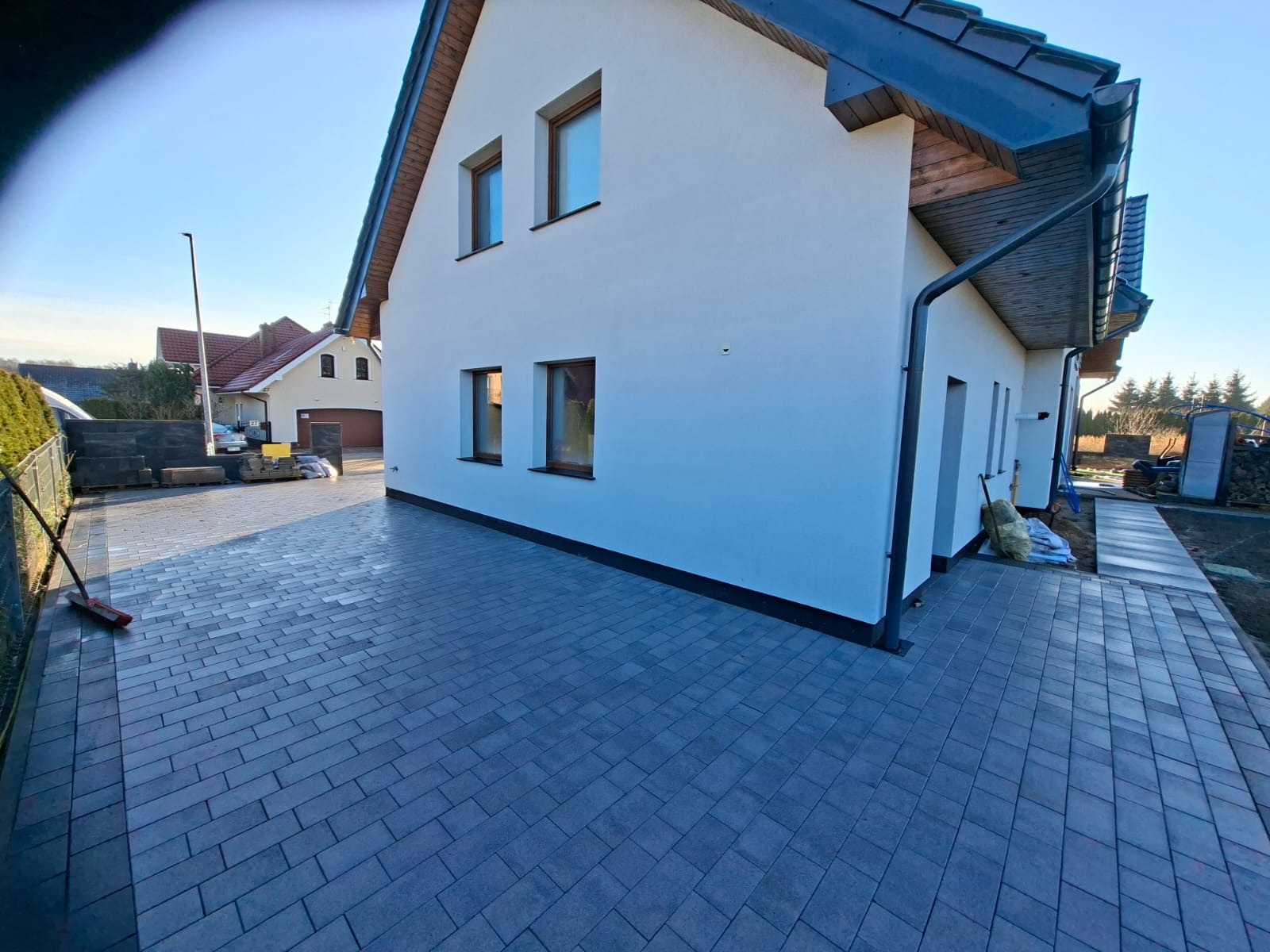 Ułożona kostka brukowa na podjeździe przed nowoczesnym domem z białymi ścianami i ciemną dachówką. Widoczne detale wykończenia i sąsiednia zabudowa.