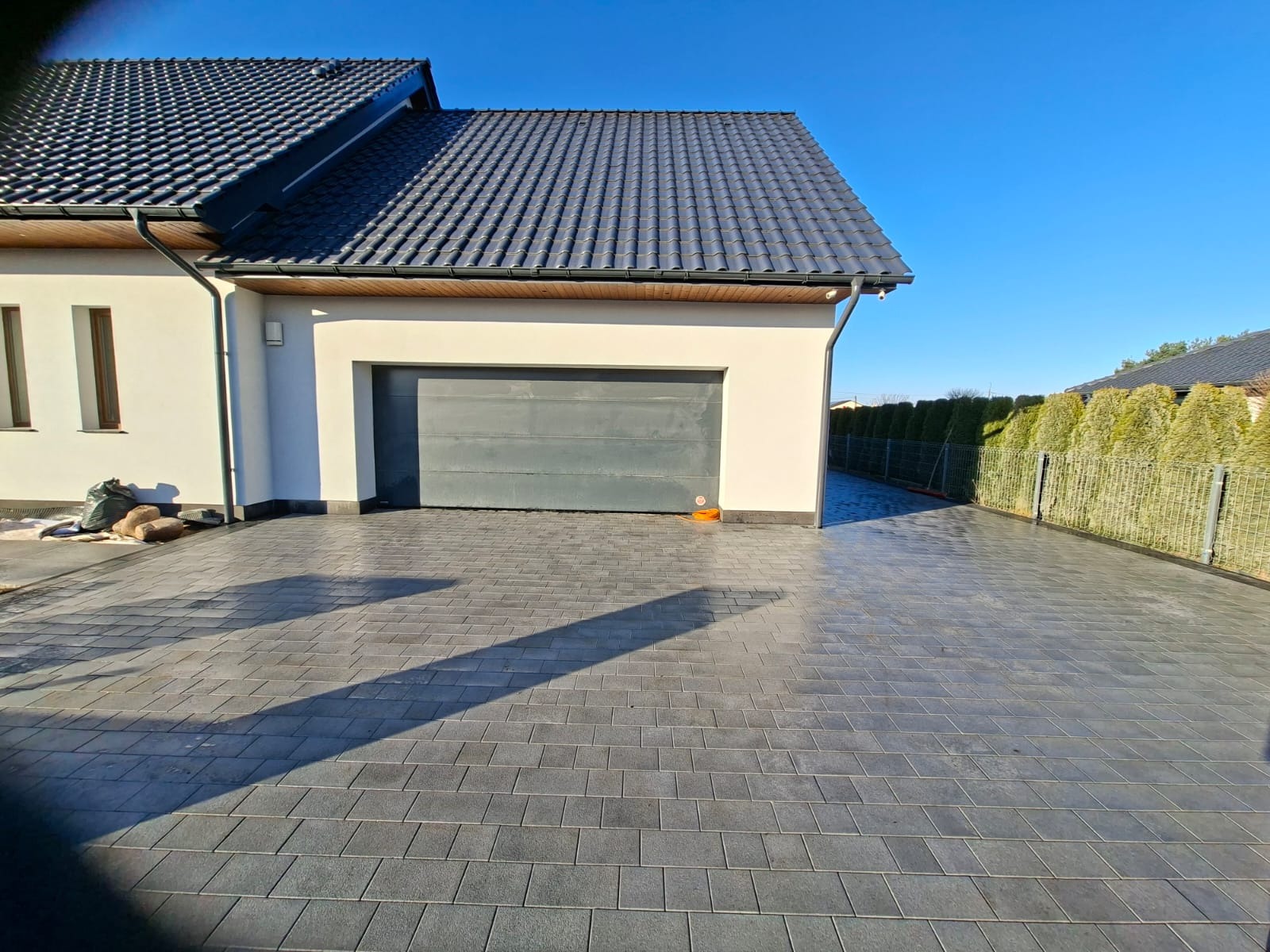 Ułożona, szara kostka brukowa na podjeździe do garażu w nowoczesnym domu z ciemną dachówką i ogrodzeniem z tuj. Widok w słoneczny dzień.