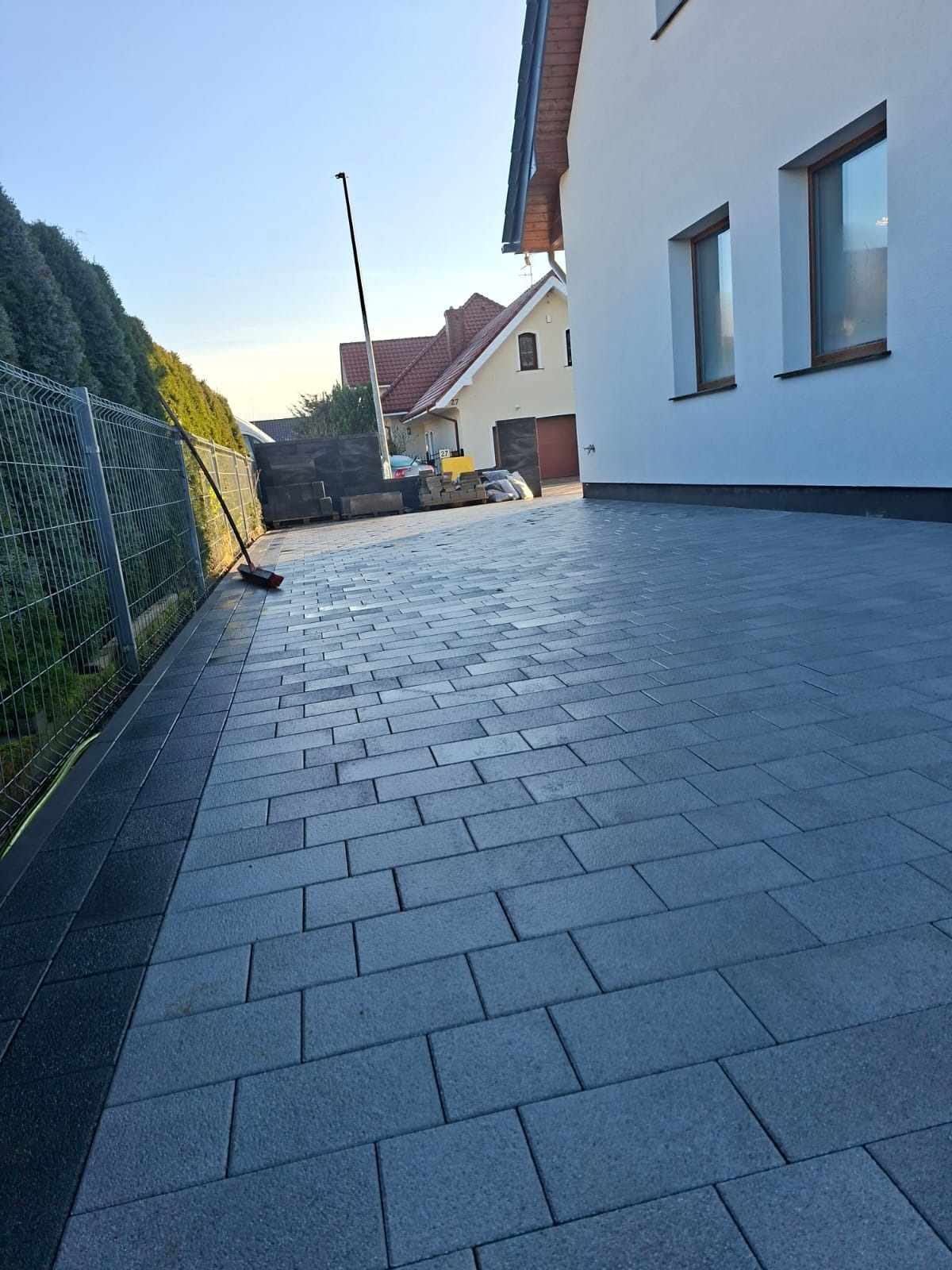 Ułożona, grafitowa kostka brukowa na podjeździe, obok ogrodzenia z siatki i budynku z białymi ścianami. W tle widoczny dom z czerwonym dachem.