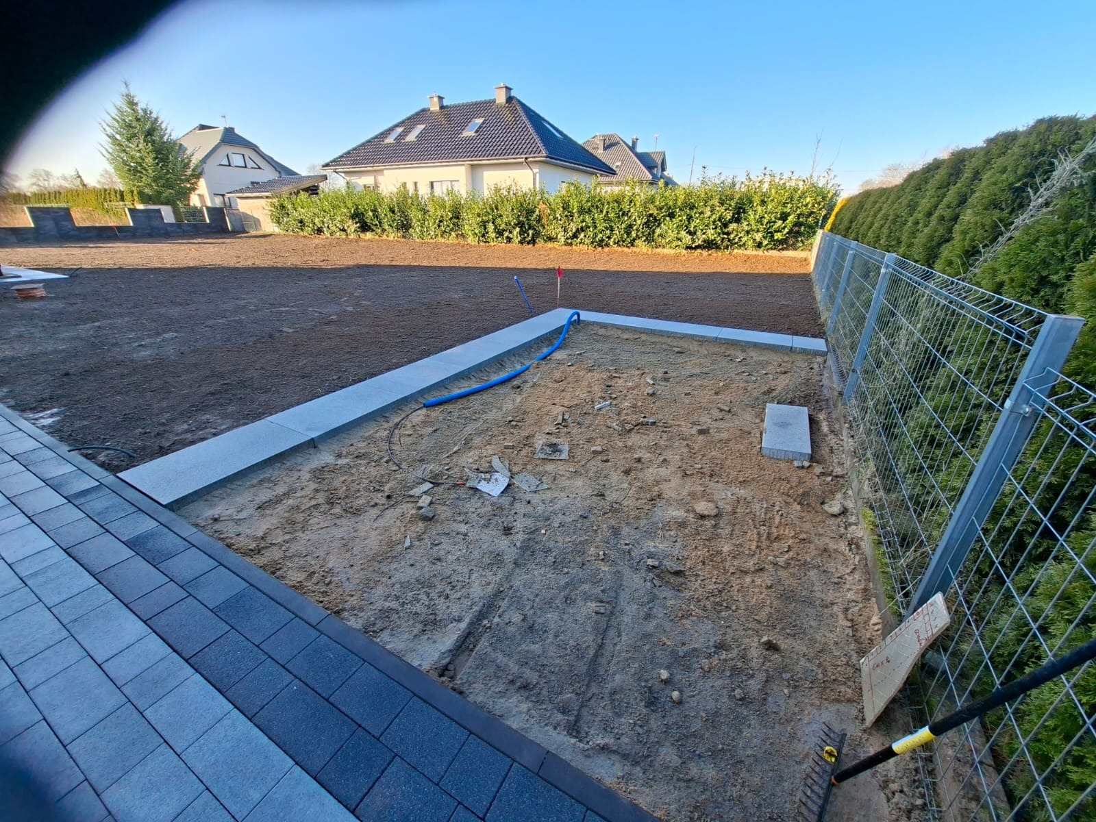 Przygotowanie terenu pod trawnik z rolki: wyrównana ziemia, obrzeża z kostki, częściowo ułożona kostka brukowa, widoczny fragment ogrodzenia i węża do nawadniania.