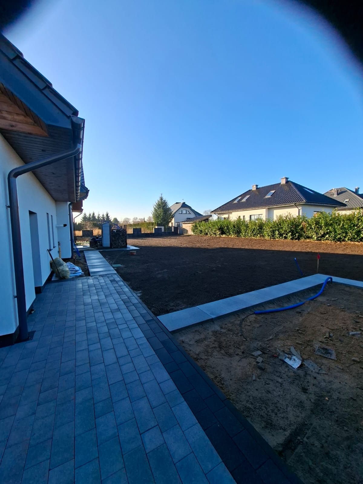Przygotowany teren pod trawnik z rolki, widoczne obrzeża i nawodnienie. Częściowo ułożona kostka brukowa przy budynku. Słoneczny dzień.