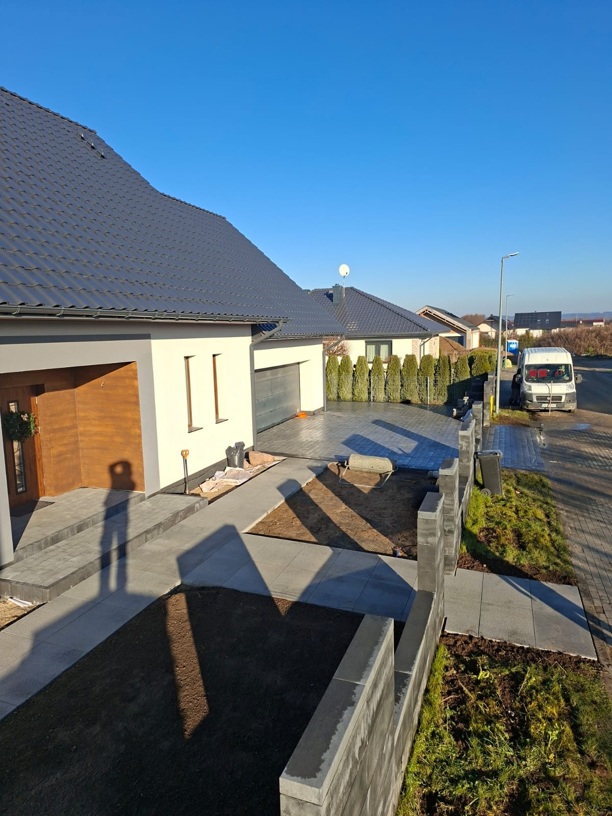 Nowoczesny dom z szarej kostki brukowej i częściowo ukończonym ogrodzeniem z betonowych bloczków. Widoczny cień osoby robiącej zdjęcie. Słoneczny dzień.