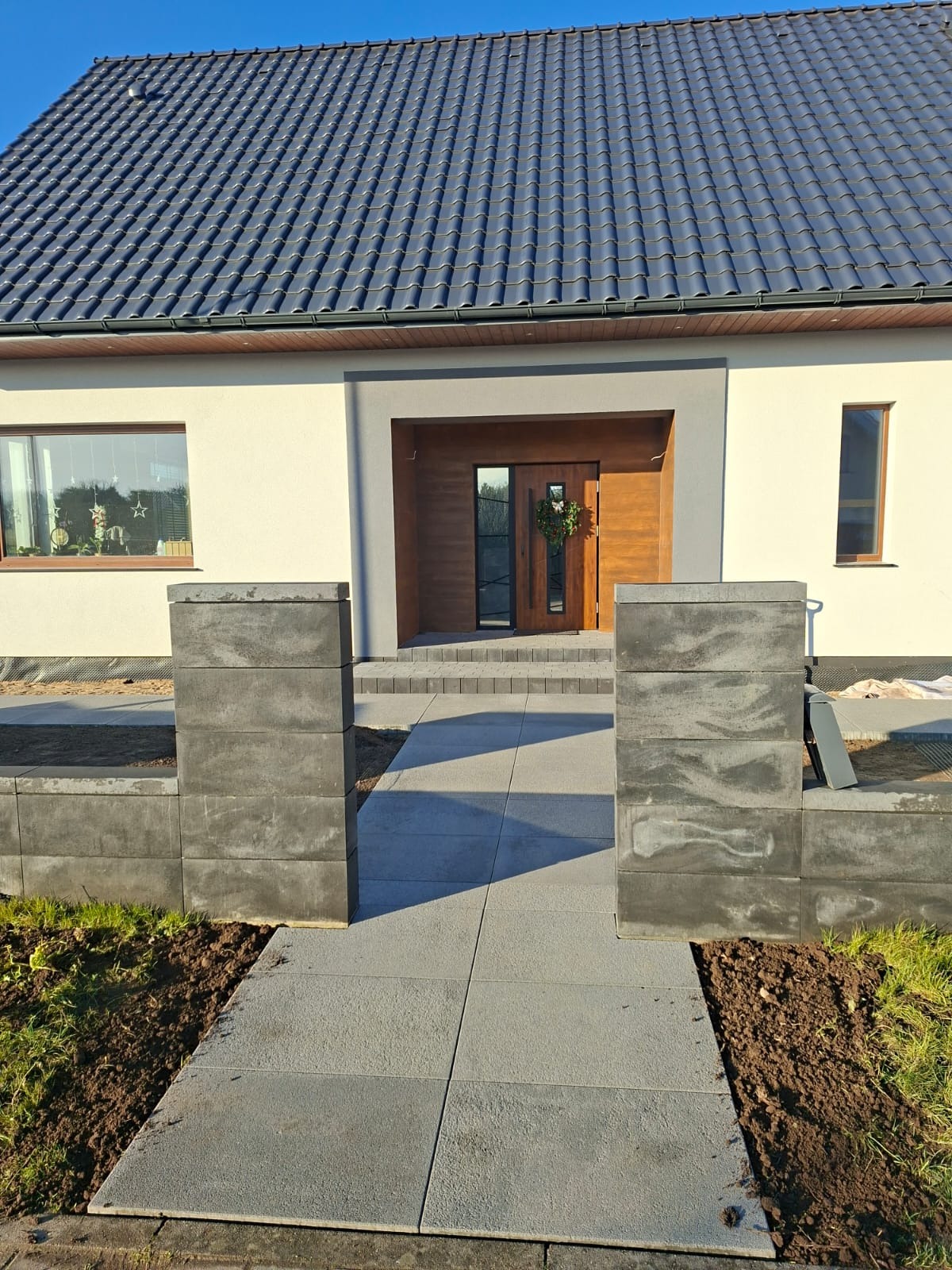 Nowoczesny dom jednorodzinny z szarą kostką brukową prowadzącą do wejścia. Widoczne elementy ogrodzenia z betonowych bloczków i świeżo obsadzona ziemia.