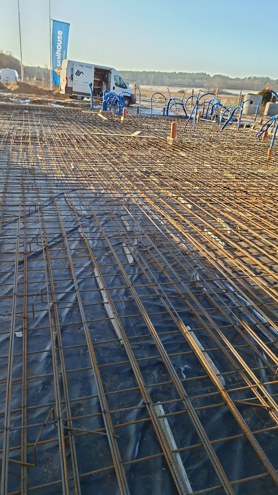 Zbrojenie fundamentów na czarnej folii, z widocznymi niebieskimi rurami instalacji i firmowym vanem w tle, w słoneczny dzień. Detal konstrukcji.