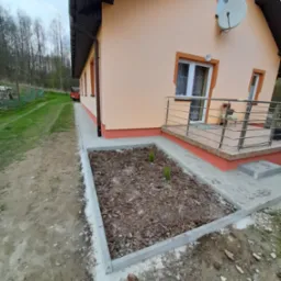 Dom jednorodzinny z ocieploną elewacją w odcieniach beżu i brązu, widoczny fragment tarasu z metalową balustradą oraz świeżo wykończona rabata z korą i młodymi sadzonkami, teren wokół w trakcie...