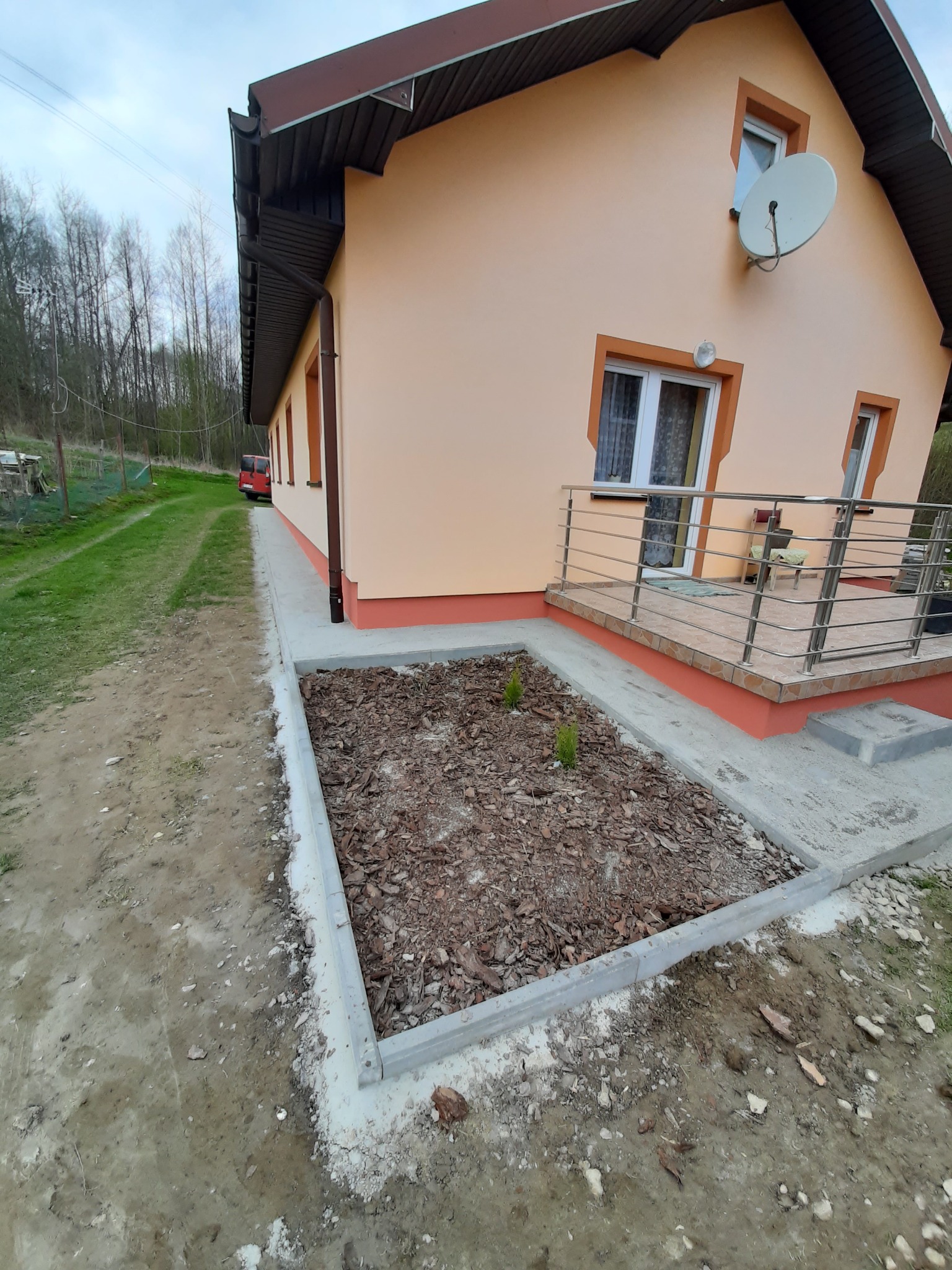 Dom jednorodzinny z ocieploną elewacją w odcieniach beżu i brązu, widoczny fragment tarasu z metalową balustradą oraz świeżo wykończona rabata z korą i młodymi sadzonkami, teren wokół w trakcie...