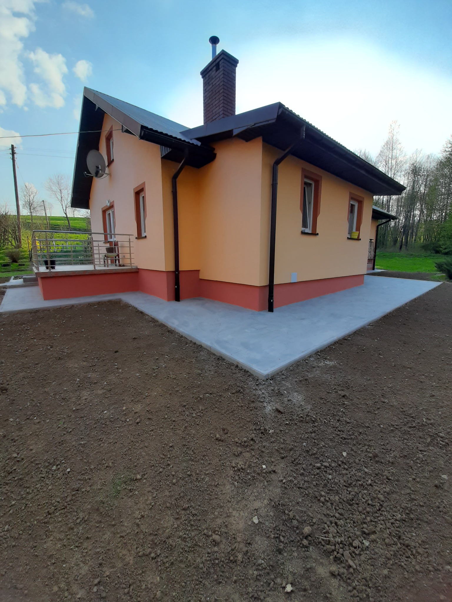 Ocieplony dom jednorodzinny z nową elewacją w odcieniach pomarańczy, z ciemnobrązowym dachem i wykończeniami, widoczny z perspektywy gruntu, z przygotowanym terenem pod trawnik.