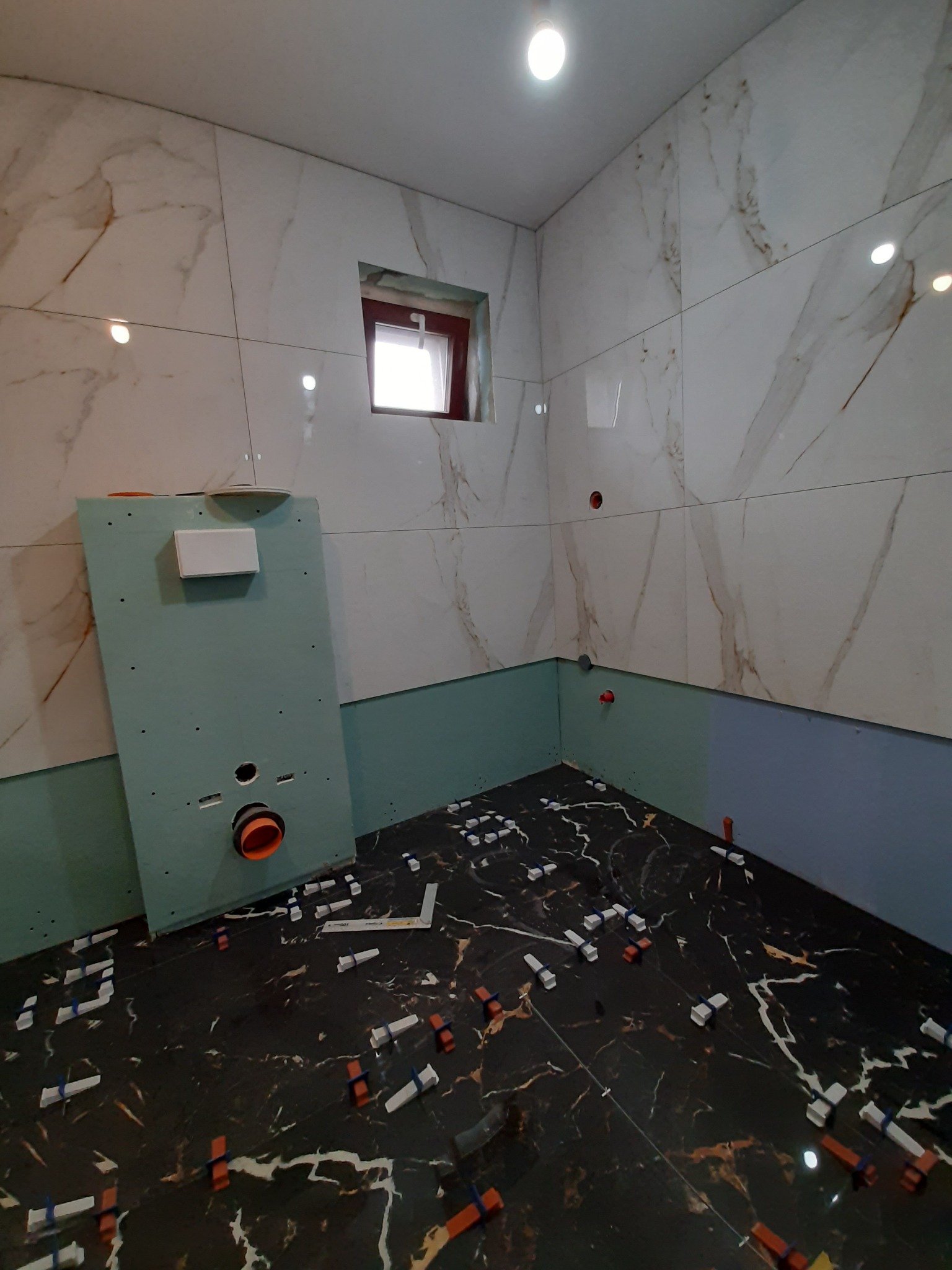 Łazienka w trakcie wykańczania: białe płytki imitujące marmur na ścianach, czarne płytki podłogowe z białymi żyłami, płyta karton gips z instalacją podtynkową, poziomica i kliny do płytek...