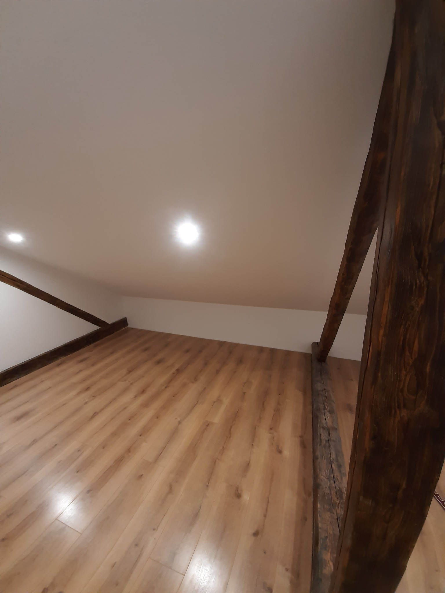 Poddasze po remoncie z drewnianą podłogą, widoczne drewniane belki konstrukcyjne i białe ściany pod skosem, oświetlone punktowo.