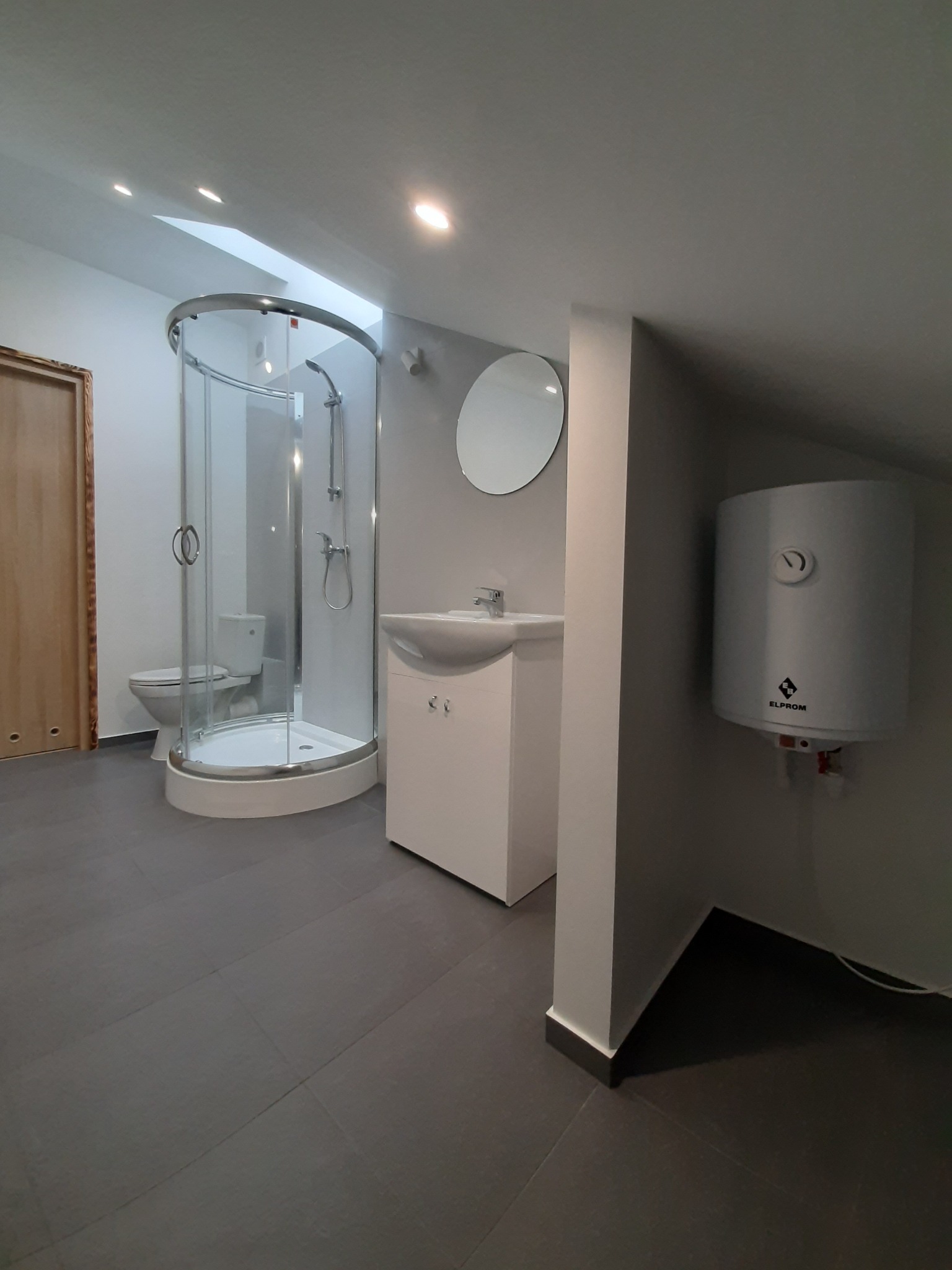 Nowoczesna łazienka na poddaszu z kabiną prysznicową, okrągłym lustrem, białą szafką pod umywalką i bojlerem Elprom.