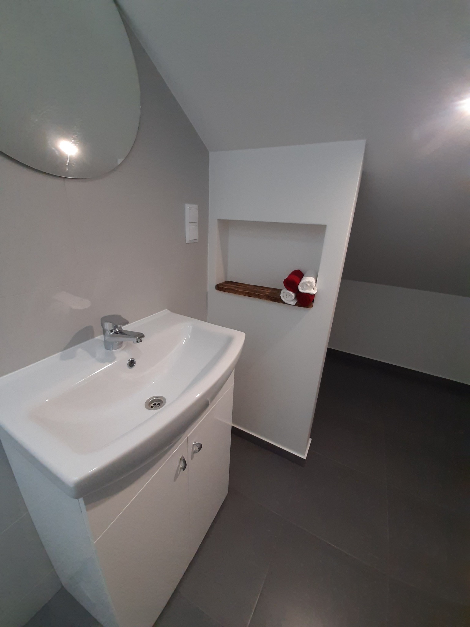 Nowoczesna łazienka na poddaszu z białą umywalką nablatową z szafką, okrągłym lustrem i wnęką z drewnianą półką z ręcznikami w kolorze czerwieni i bieli.