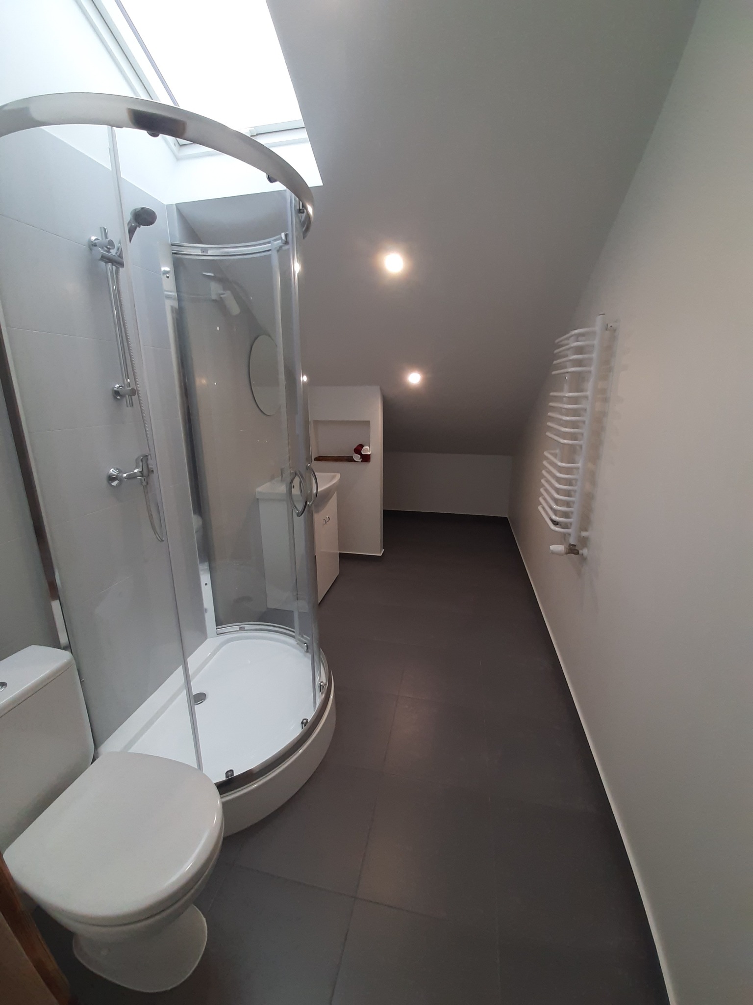 Nowoczesna łazienka na poddaszu z kabiną prysznicową z zaokrąglonymi szybami, wiszącą toaletą, grzejnikiem drabinkowym i oknem dachowym.