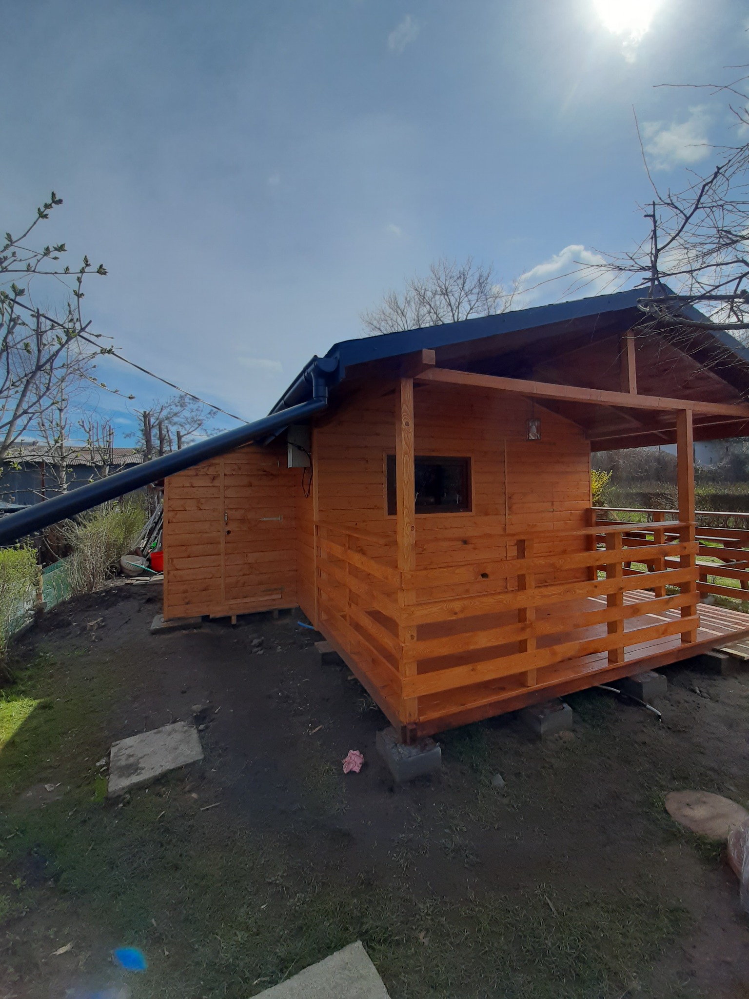 Nowo wybudowany, drewniany domek letniskowy z zadaszonym tarasem, widoczny z zewnątrz, z rynną spustową i elementami instalacji elektrycznej na ścianie.