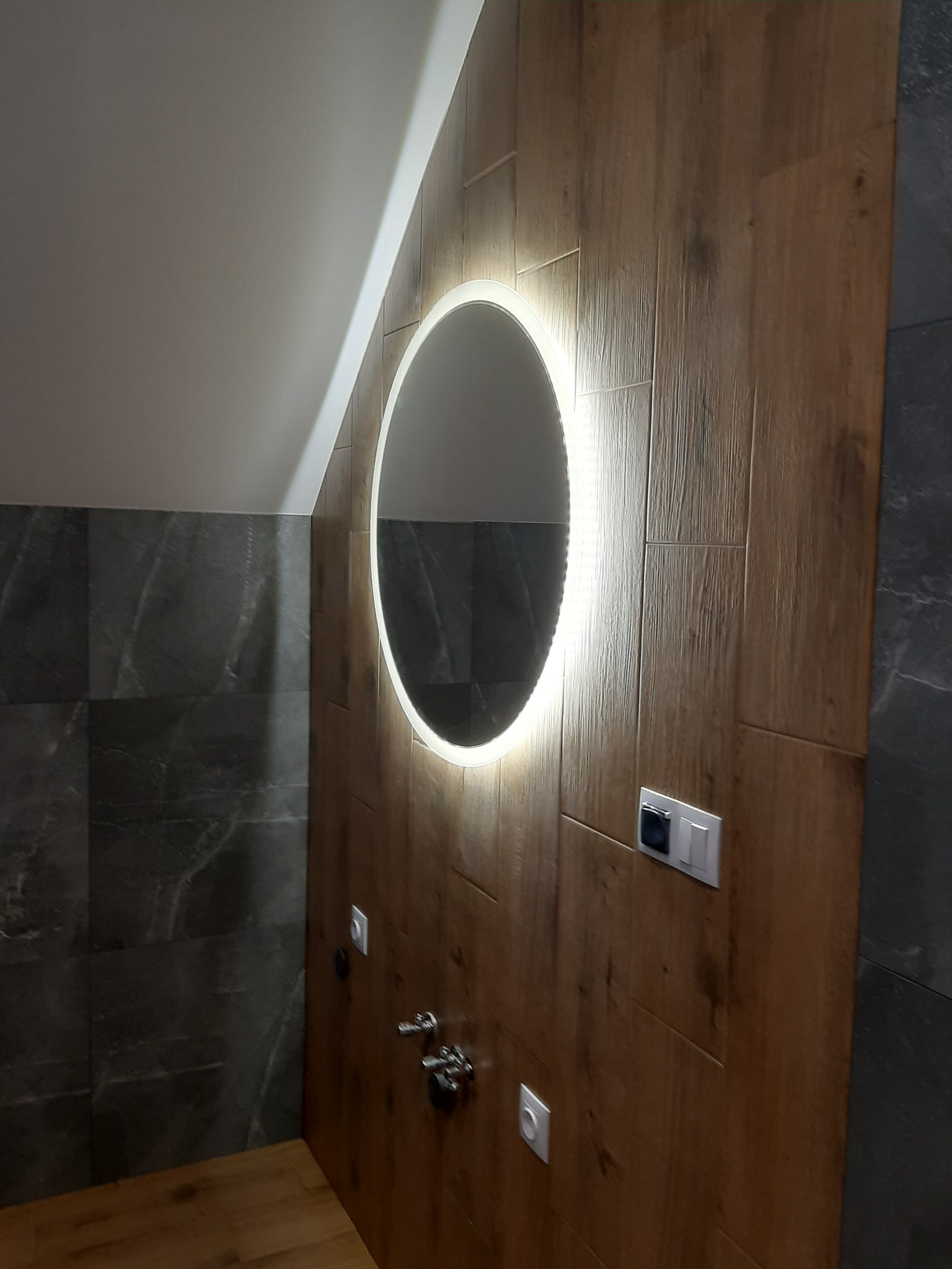 Nowoczesna łazienka na poddaszu z okrągłym lustrem podświetlanym LED, ściany wyłożone płytkami imitującymi drewno i szarym marmurem.