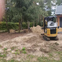 Żółta minikoparka CAT 301.5 na terenie budowy, z widocznymi hałdami ziemi i kamieni, w tle drzewa i fragment domu z drewnianą elewacją.