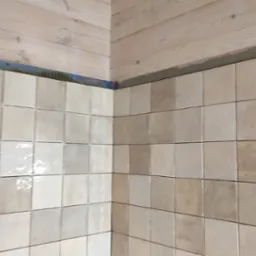Ściana wyłożona beżowymi, kwadratowymi płytkami ceramicznymi, widoczne drewniane wykończenie sufitu i górnej krawędzi ściany, świeżo położone płytki z widocznymi śladami zaprawy.