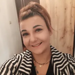 Uśmiechnięta kobieta w czarno-białej marynarce w paski, z upiętymi rudymi włosami i delikatnym makijażem, pozuje na tle drewnianej szafy.