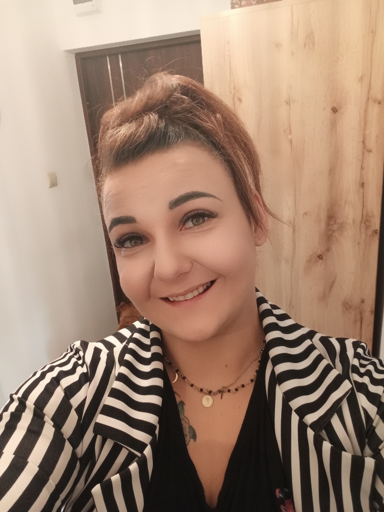Uśmiechnięta kobieta w czarno-białej marynarce w paski, z upiętymi rudymi włosami i delikatnym makijażem, pozuje na tle drewnianej szafy.