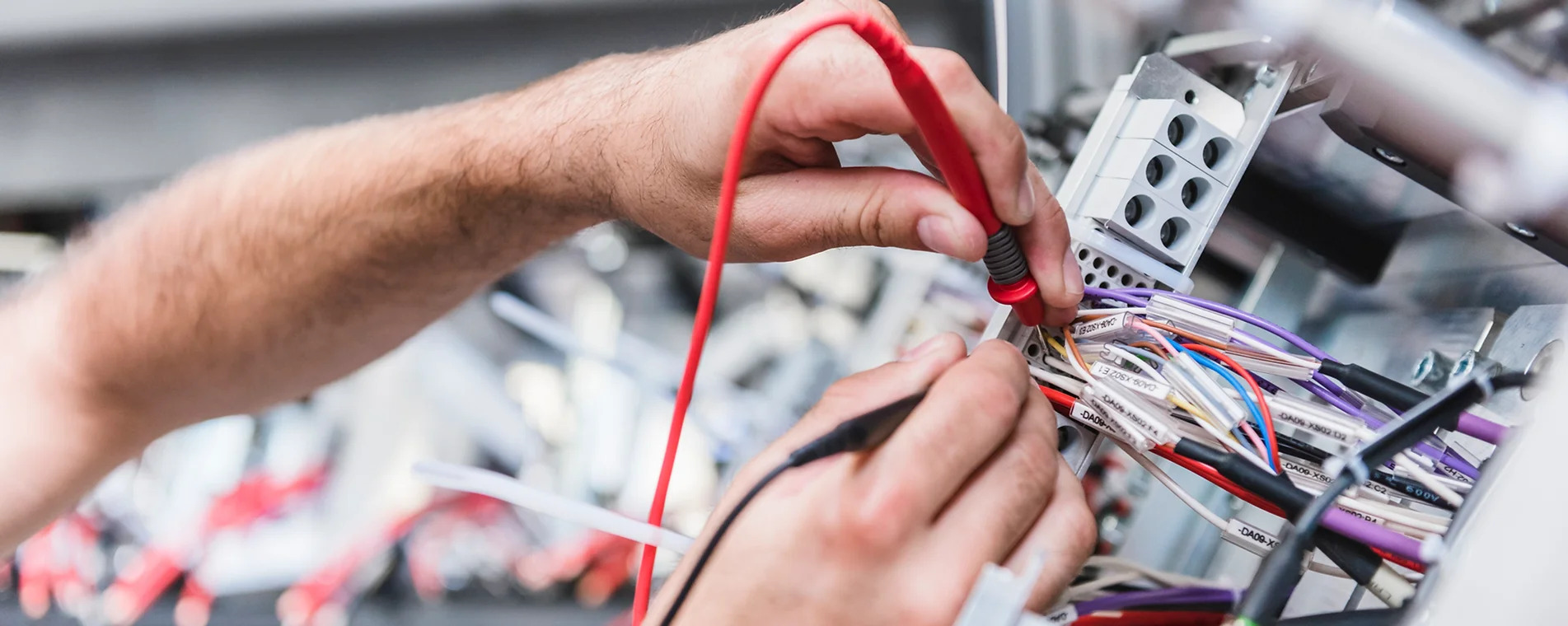 Elektryk ze Świdnicy mierzy napięcie w skrzynce rozdzielczej, używając multimetru z czerwonym i czarnym przewodem pomiarowym. Widoczne są liczne przewody z oznaczeniami, wpięte w listwę zaciskową.