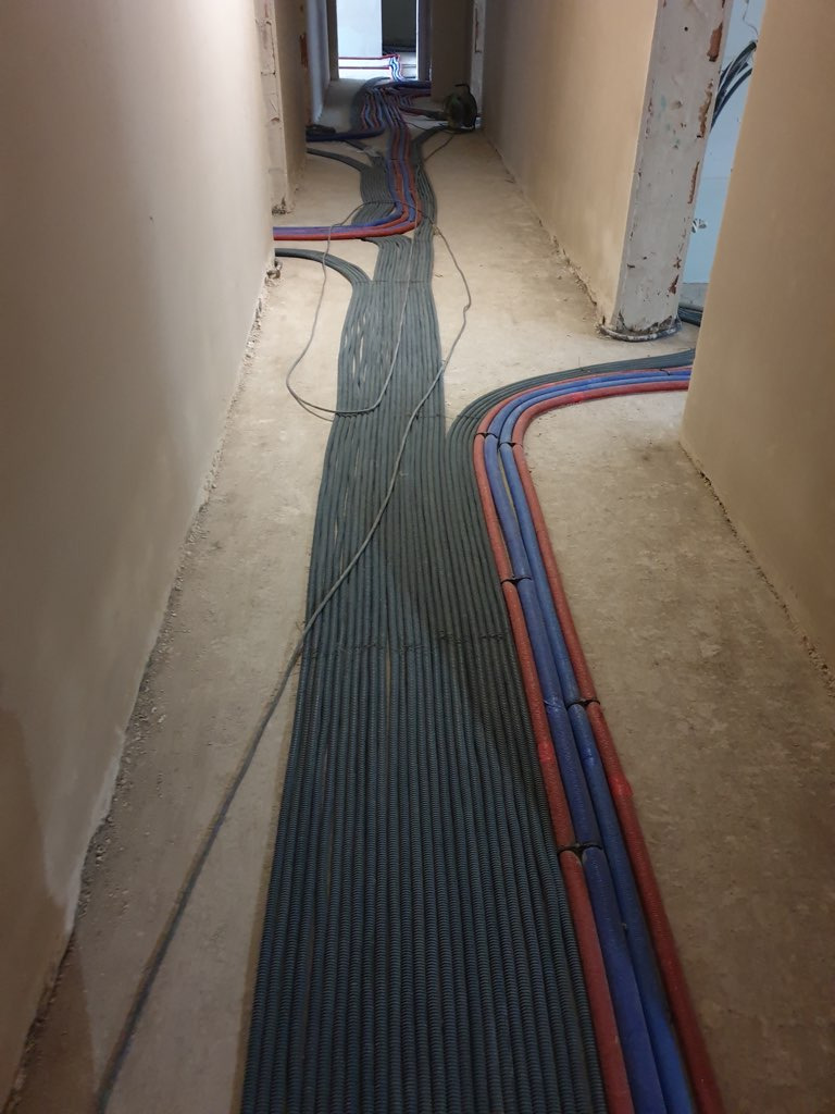 Korytarz w trakcie remontu z ułożonymi rurami instalacyjnymi w peszlach: szare, oraz czerwono-niebieskie, prowadzące wzdłuż ścian.