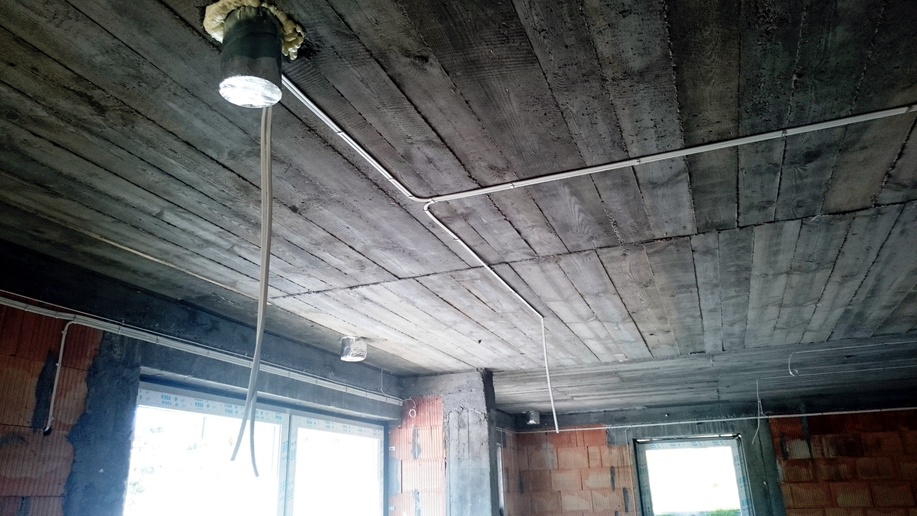 Surowy betonowy strop z widocznymi śladami szalunku i poprowadzonymi przewodami elektrycznymi w białych listwach, oczekujące na dalsze prace wykończeniowe, widoczne okna.